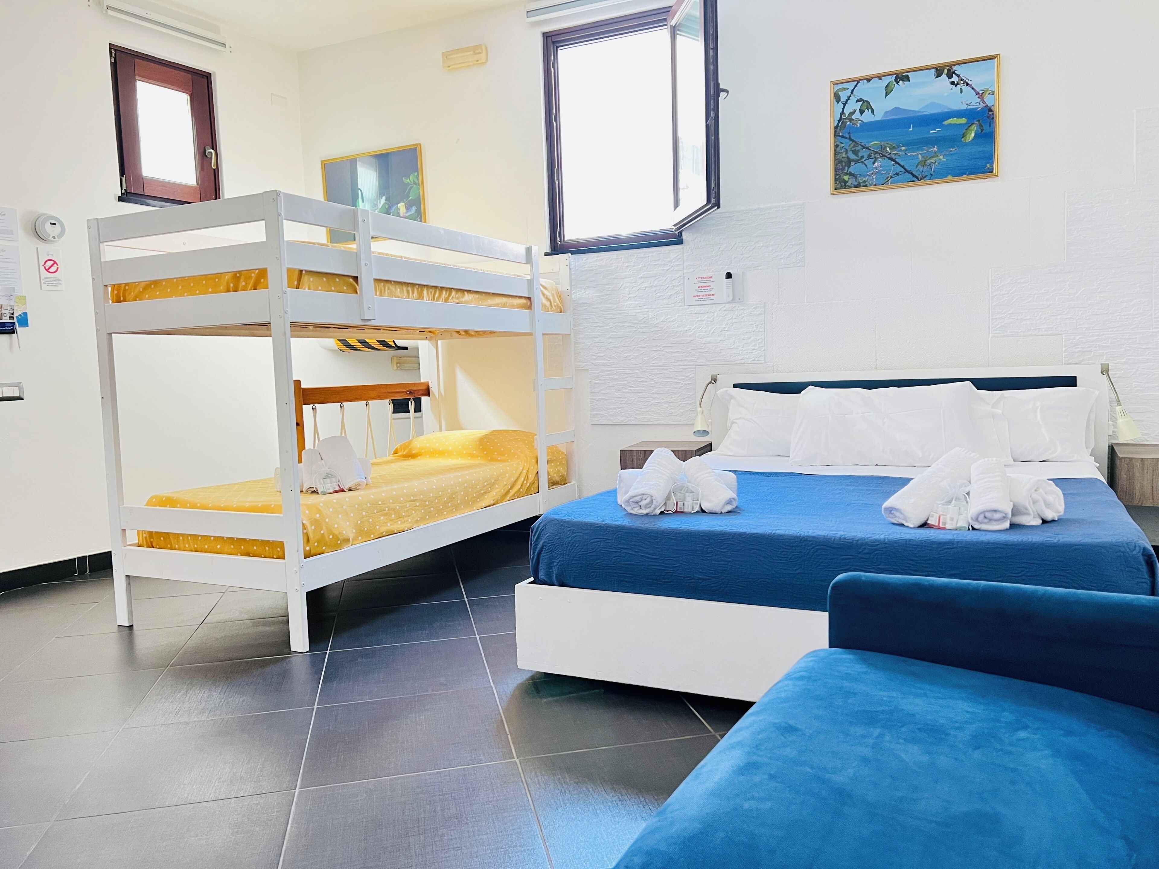 A room with a bunk bed, a single bed with white pillows, a blue bedspread, and a painting on the wall.