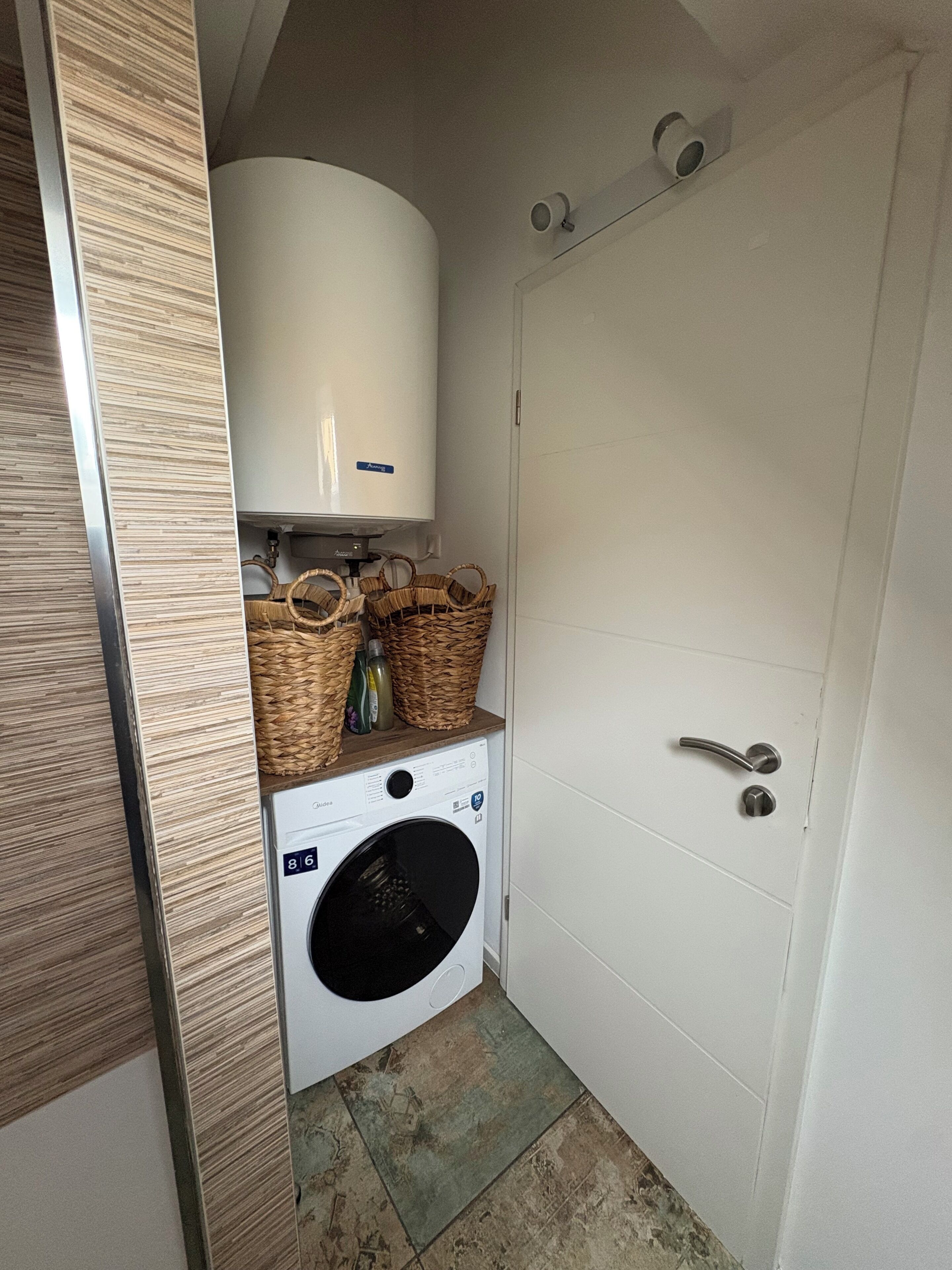 A compact laundry area with a washing machine, a water heater, and wicker baskets on a shelf.