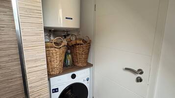 A compact laundry area with a washing machine, a water heater, and wicker baskets on a shelf.