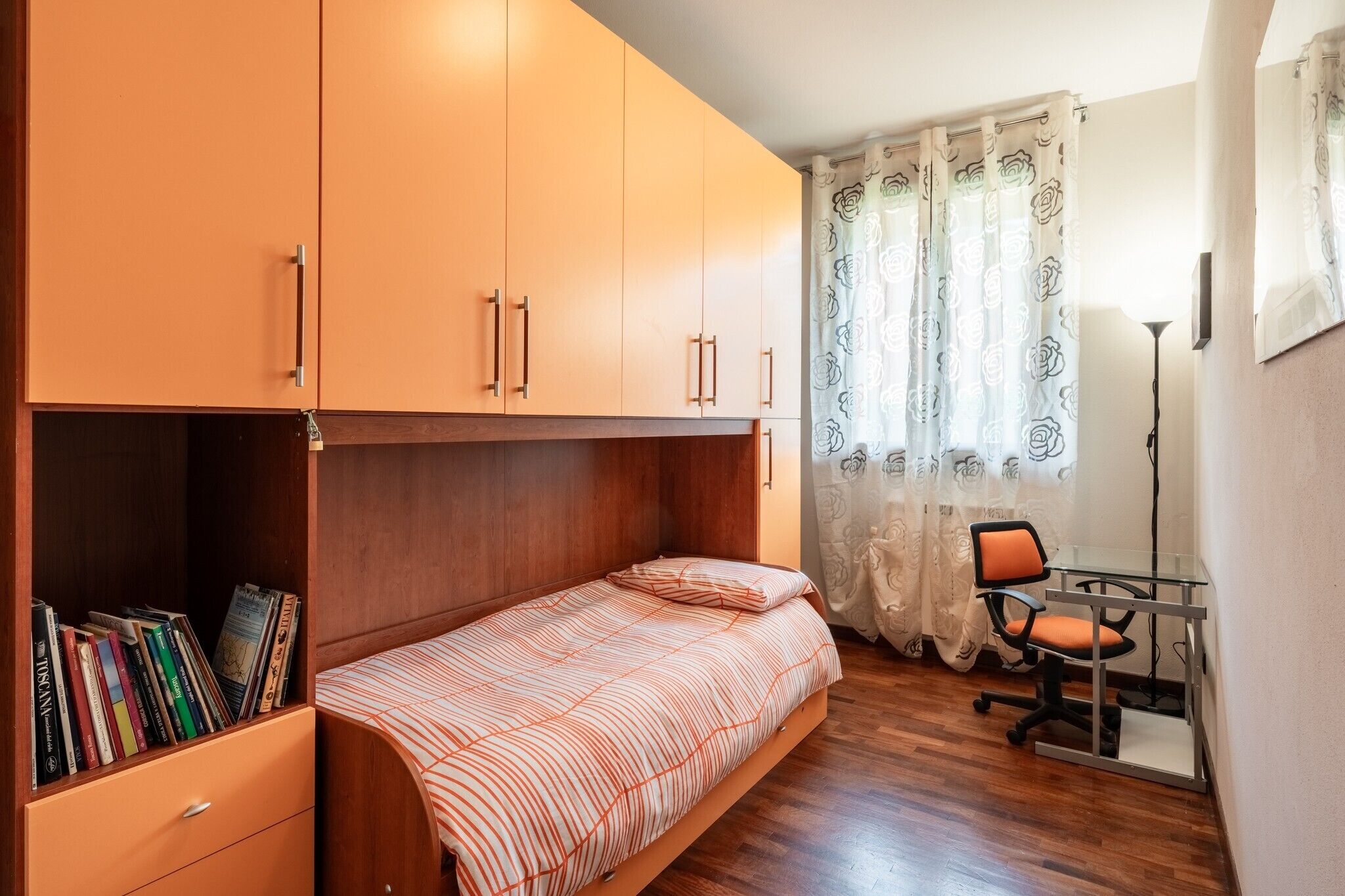 Une chambre avec un lit superposé, un bureau avec une chaise, une bibliothèque et une fenêtre ornée de rideaux à motifs floraux.