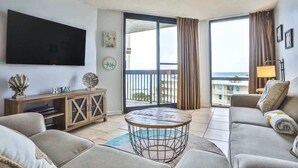 Una sala de estar con televisor de pantalla plana, un sofá, una mesa de centro y vistas al mar desde un balcón.
