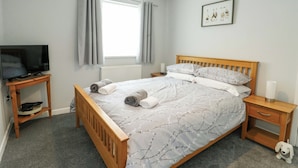 Une chambre à coucher avec un lit, un téléviseur posé sur un meuble, une table de chevet, une petite table, un tableau encadré au mur et une fenêtre avec des rideaux.