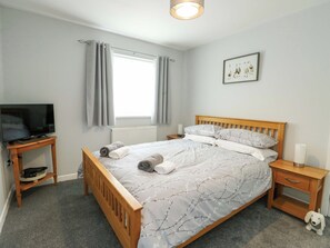A bedroom with a bed, a TV on a stand, a nightstand, a small table, a framed picture on the wall, and a window with curtains. - Min Y Cae (Cemaes Bay)