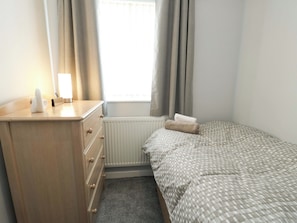 A bedroom with a bed, a dresser, a radiator, and a window with curtains. - Min Y Cae (Cemaes Bay)