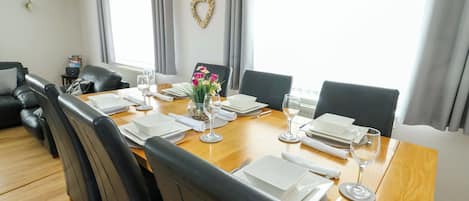Uma sala de jantar com mesa de madeira posta para seis pessoas, cadeiras de couro preto, um vaso de flores e uma decoração em formato de coração na parede.