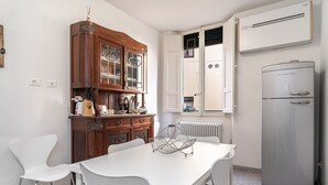 Eine moderne Küche mit einem weißen Tisch, zwei weißen Stühlen, einem Holzschrank mit Glastüren, einem eingebauten Kühlschrank und einem Fenster mit geöffneten Rollläden.