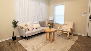 Ein Wohnzimmer mit einer Couch, einem Schaukelstuhl, einem kleinen Tisch, einer Topfpflanze und einem Fenster mit Vorhängen.