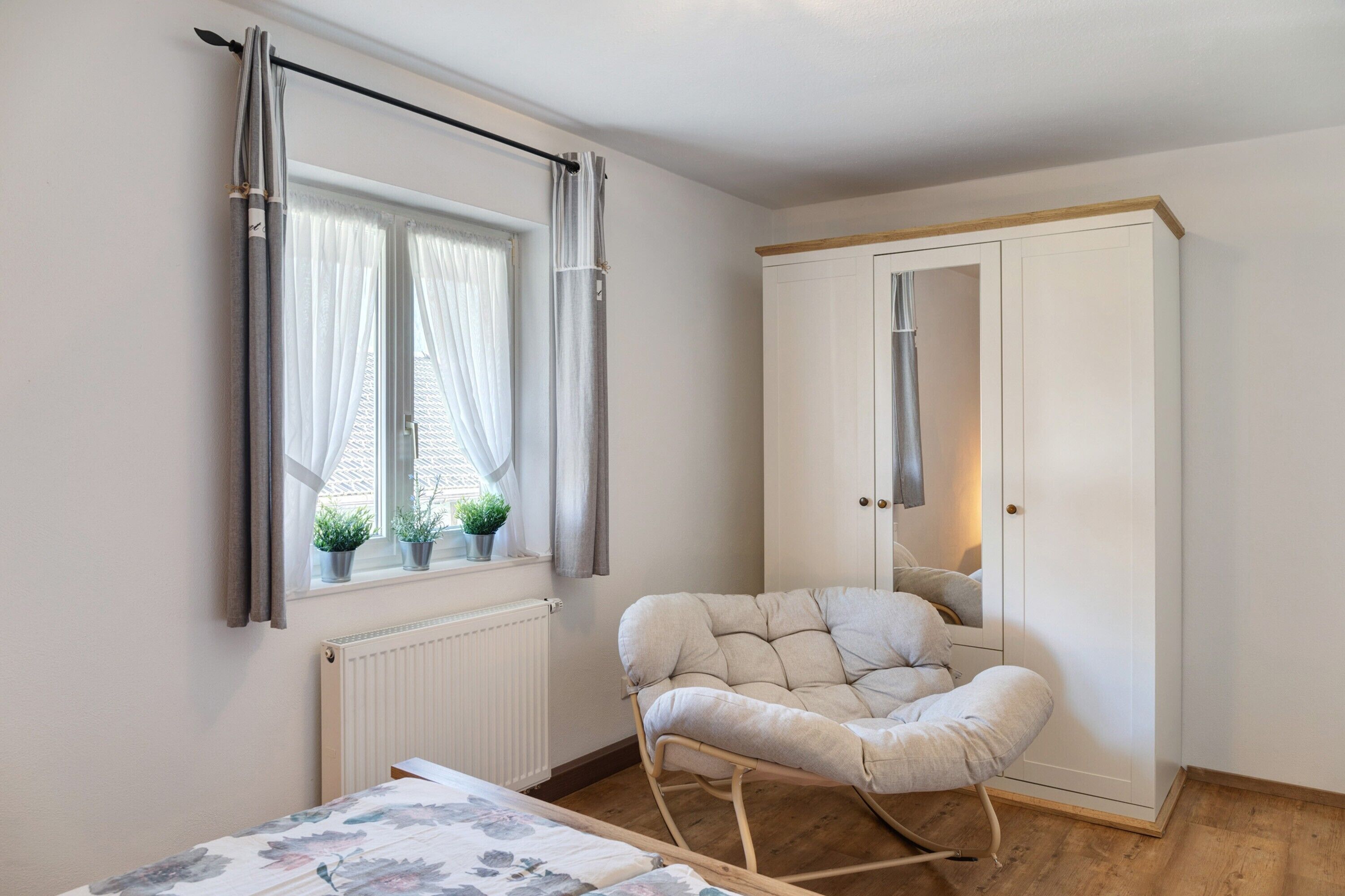 A bedroom with a bed, a rocking chair, a wardrobe, a radiator, and a window with curtains.