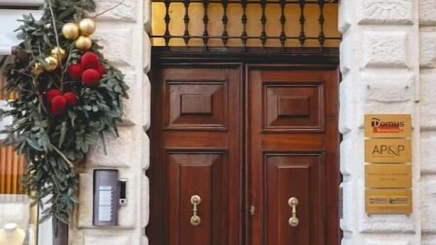 A double wooden door with a number '5' above it, a plaque with 'Dom's' and 'AP&P' text, and a wreath with red and gold decorations on the left. - Exclusive suite with Cathedral view | Elegant design in the heart of Ferrara (Ferrara)