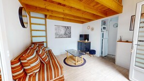 Ein gemütliches Zimmer mit Holzbalkendecke, einer Couch mit gestreiften Kissen, einem kleinen Tisch mit einem Drahtkorb, einem Fernseher und einem Kühlschrank.