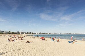 Ein Strand mit badenden und sonnenden Menschen, ein Steg im Hintergrund und Gebäude entlang der Küste.