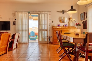 A dining area with wooden chairs, a wooden table, a television, and a view through a glass door to an outdoor seating area. - Casa Saudade, a Home in Madeira (Porto Santo)