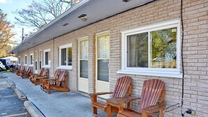 A row of wooden Adirondack chairs outside a brick building with multiple windows.