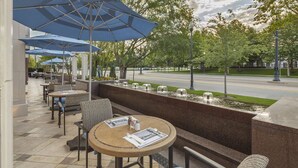 An outdoor patio with round tables and blue umbrellas, overlooking a road and greenery. - Garden 1 King, Little America, Unwind and Relax, Near Temple Square (Salt Lake City)
