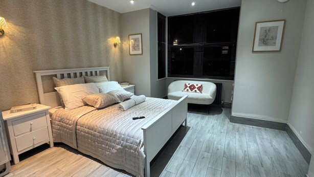 Un dormitorio con cama, mesitas de noche, una silla, una ventana con persianas y un reloj de pared.