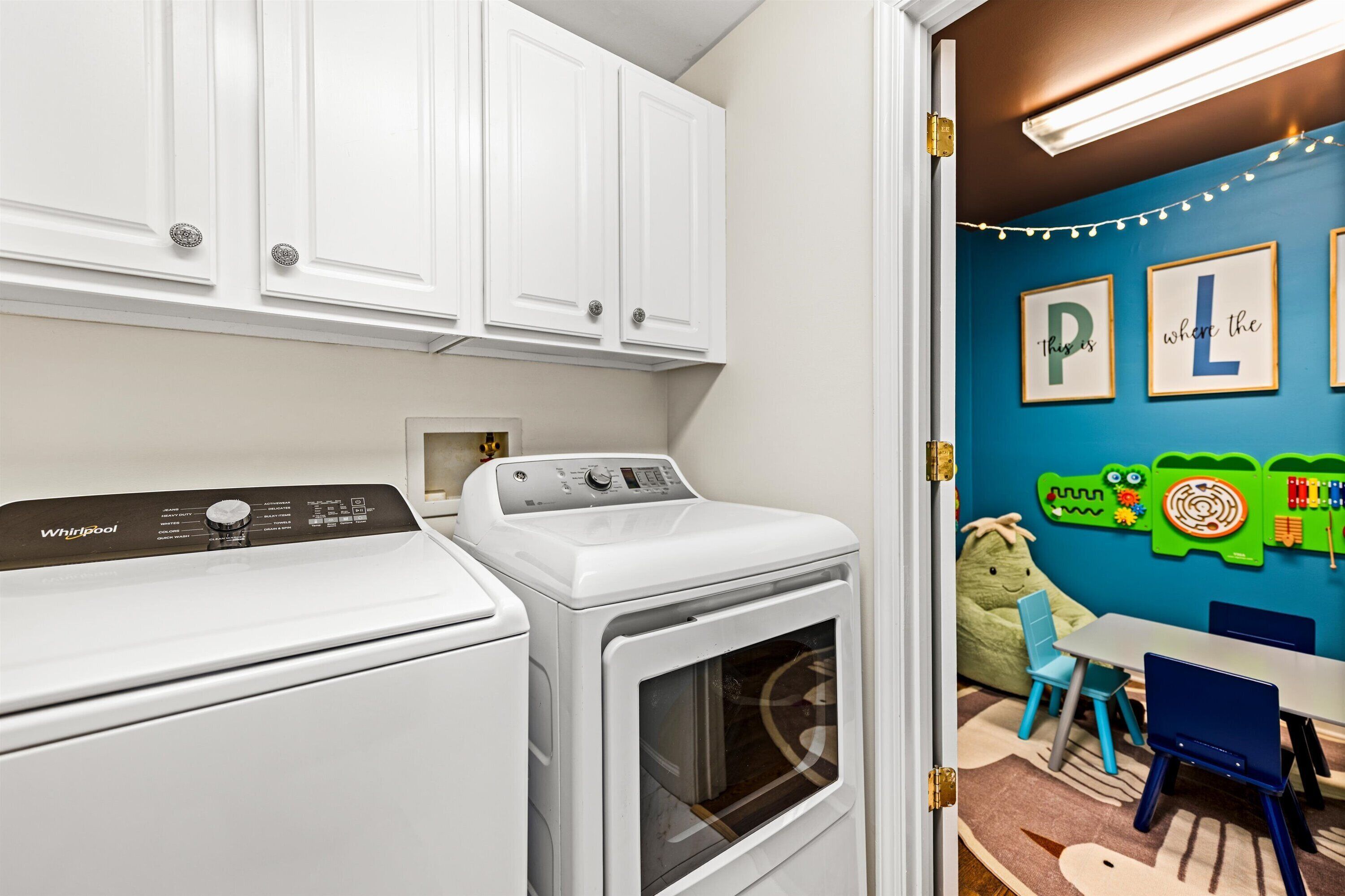 A laundry room with a Whirlpool washer and dryer, a blue wall with children's artwork, and a small table with chairs.