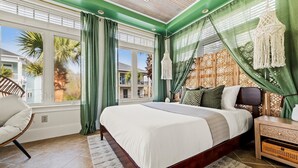A bedroom with a large bed, a chair, a nightstand, and a view of palm trees outside. - Walk 2 Beach |Theater, Games, Putt, Pool, Sleep 22 (North Myrtle Beach)