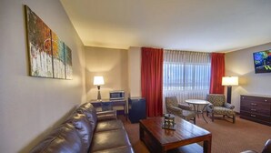 Camera d'albergo con divano in pelle, tavolino in legno, due poltrone, televisore, scrivania con lampada e un quadro di grandi dimensioni.
