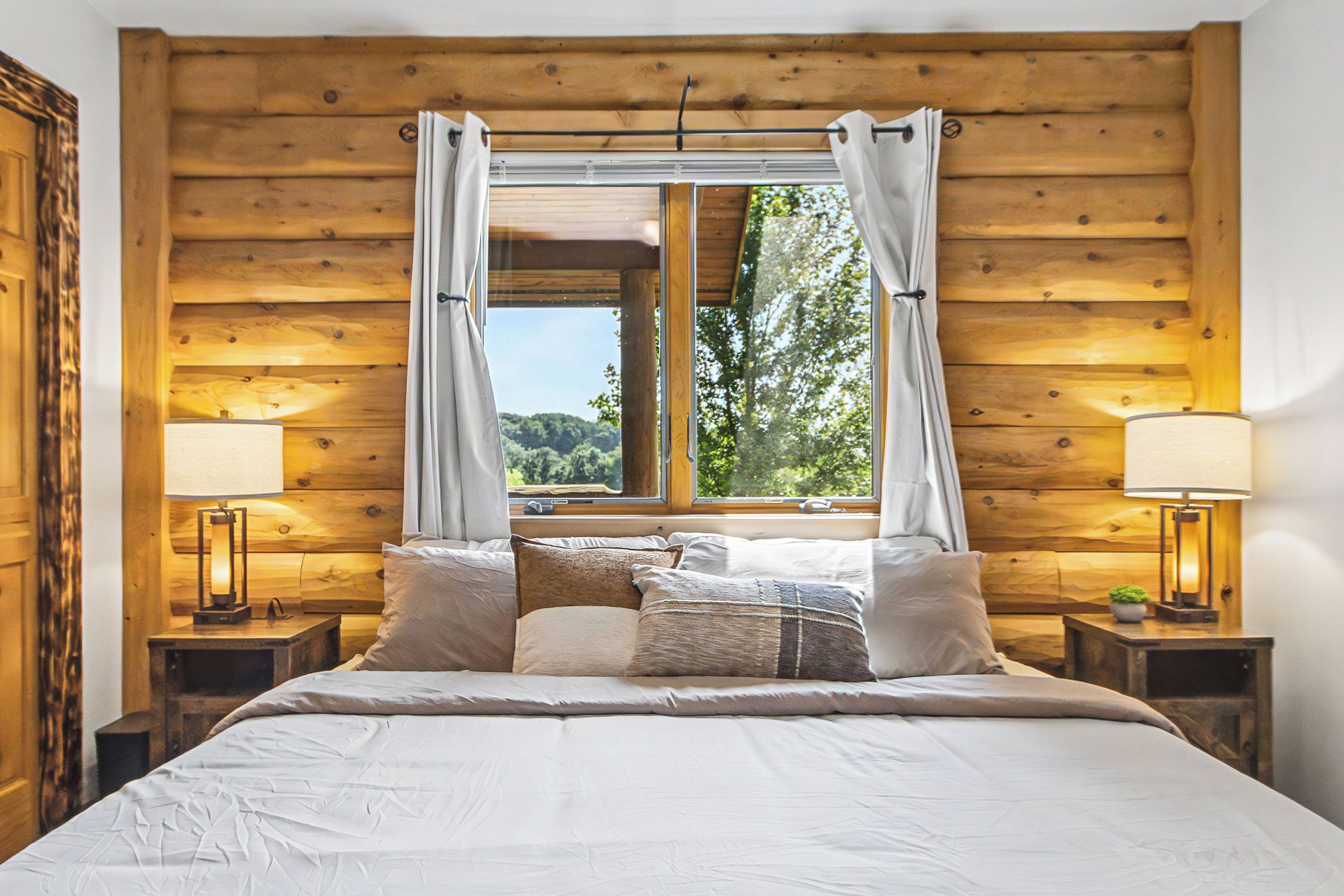 A bedroom with a wooden log wall, a large bed with white bedding, two bedside tables with lamps, and a window with curtains.