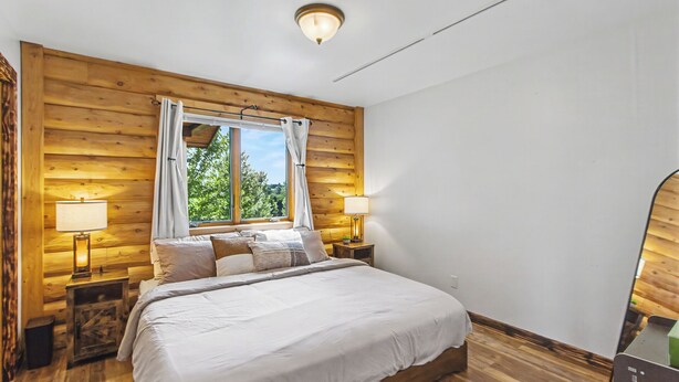 Un dormitorio con cabecera de madera, un ventanal grande, una cama con ropa blanca, dos lámparas de noche y una mesita de noche con lámpara.
