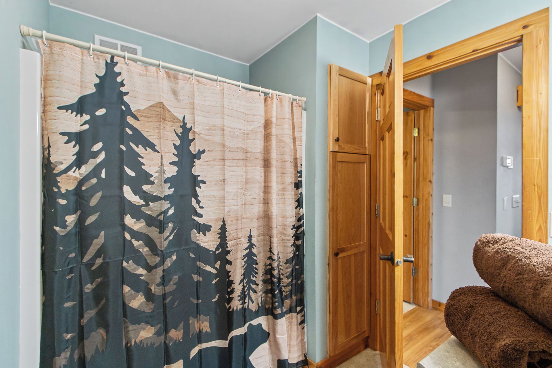Una habitación con cortina de ducha con estampado de bosque, una puerta de madera y una silla marrón.