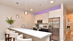 A modern kitchen with a central island, bar stools, stainless steel appliances, and decorative wall art. - New Condo Rehoboth 3BR w/4 E-Bikes, Pool + Gym (Rehoboth Beach)