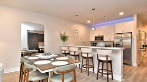 A modern kitchen with a central island, bar stools, and a dining table with placemats. - New Condo Rehoboth 3BR w/4 E-Bikes, Pool + Gym (Rehoboth Beach)
