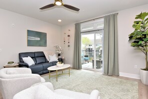 A modern living room with a sofa, armchairs, a coffee table, a ceiling fan, and a large window with curtains. - New Condo Rehoboth 3BR w/4 E-Bikes, Pool + Gym (Rehoboth Beach)