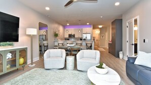 A modern living room with a sofa, armchairs, a coffee table, a TV, a ceiling fan, and a kitchen in the background. - New Condo Rehoboth 3BR w/4 E-Bikes, Pool + Gym (Rehoboth Beach)