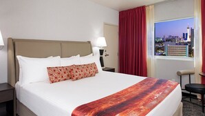 Habitación de hotel con cama, mesita de noche, lámpara, silla y ventana con vista a la ciudad.