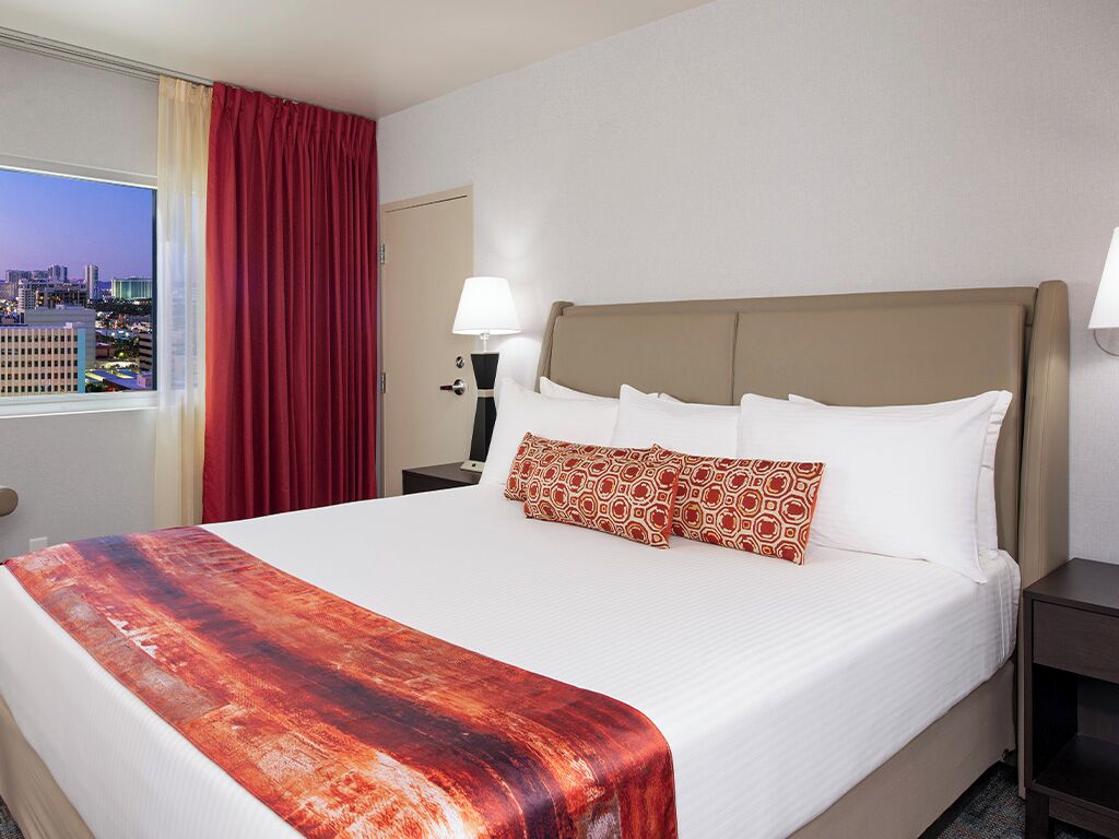 A hotel room with a large bed, a window with a city view, a red curtain, and a nightstand with a lamp.
