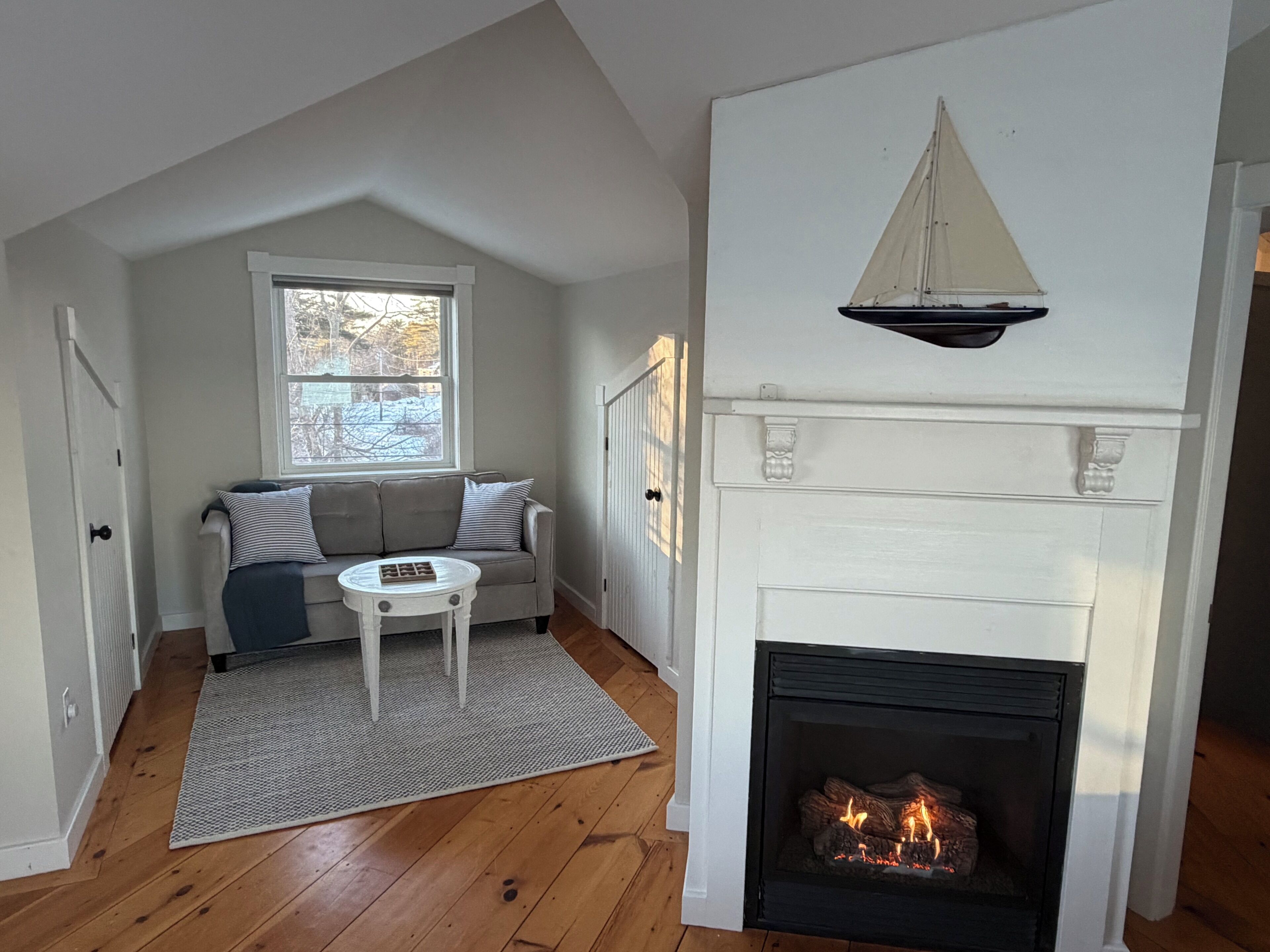 A room with a fireplace, a sofa, a small table, and a window with a view of trees.