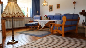 Un salon avec un canapé bleu, une chaise en bois, une table recouverte d’une nappe en dentelle, un lampadaire et un luminaire suspendu.