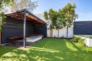 Un patio trasero con una pérgola de madera, césped y valla blanca.