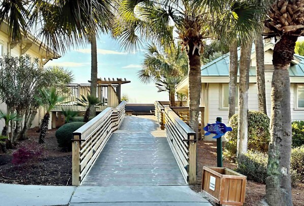 A wooden boardwalk leading to a building with a pergola, surrounded by palm trees and a signpost.