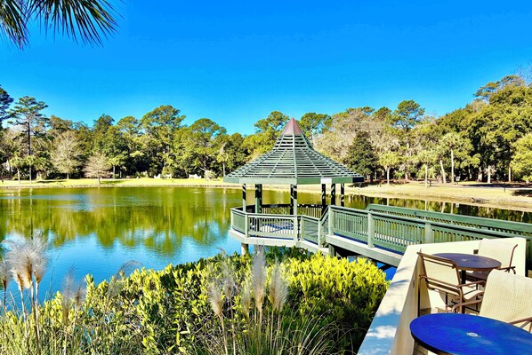 A gazebo with a green roof and white railing, overlooking a calm lake, surrounded by lush greenery and a clear blue sky.