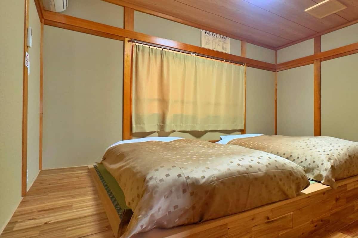 A room with a wooden bed frame, two beds, a window with a curtain, and a ceiling fan.