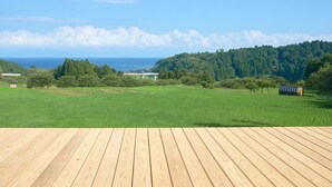 A wooden deck with a view of a green field, trees, and a distant body of water. - Yuushinan (Nanao)