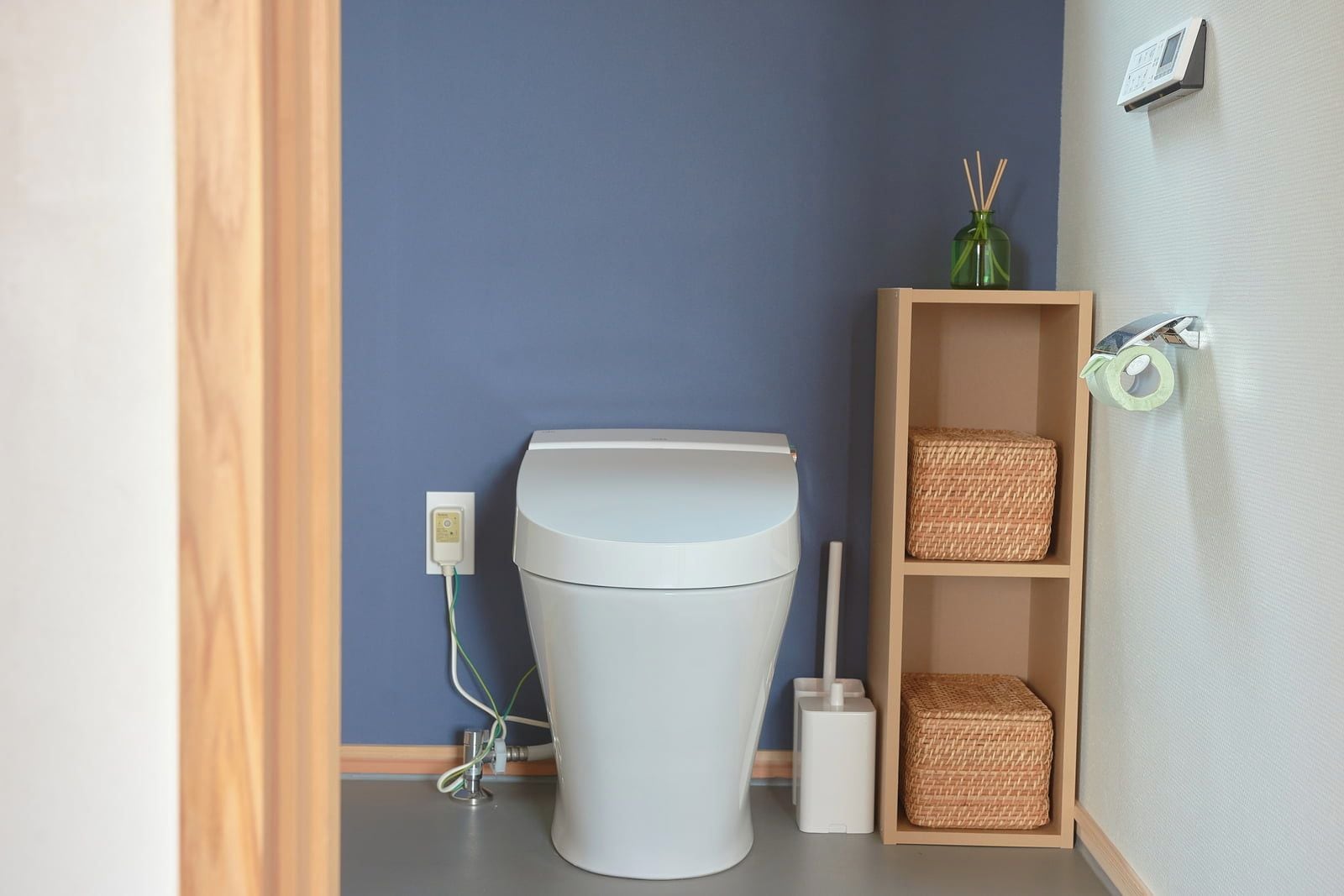 A modern bathroom with a white toilet, a wall-mounted bidet, a shelf with baskets, and a power outlet.