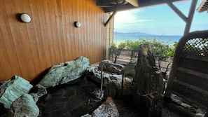 A wooden interior with a stone hot tub, wooden walls, and a view of the sea. - Ryokan Hama no Ya (Kyotango)