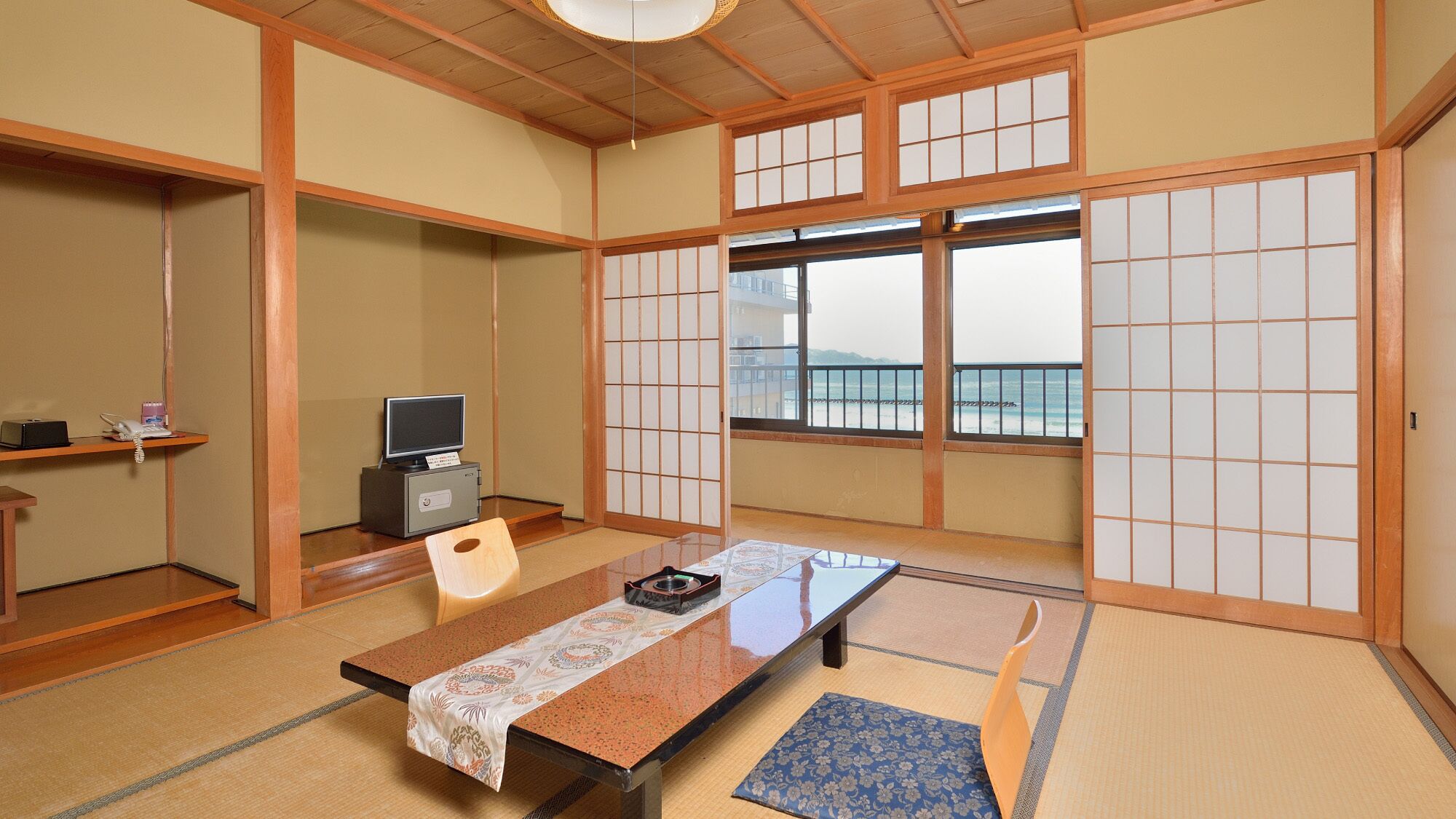 A traditional Japanese room with tatami flooring, a low table, a television, and a view of the sea through large windows.