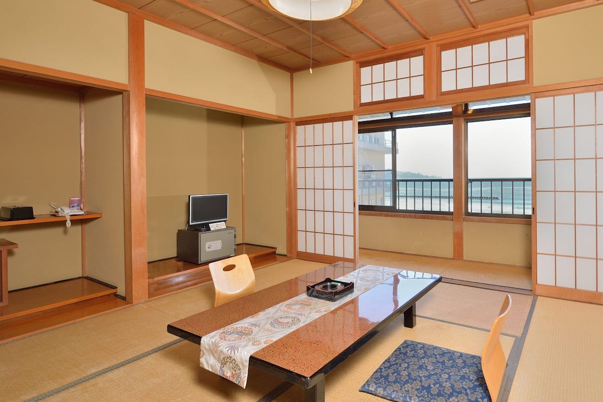 A traditional Japanese room with tatami flooring, a low table, a television, and a view of the sea through large windows.