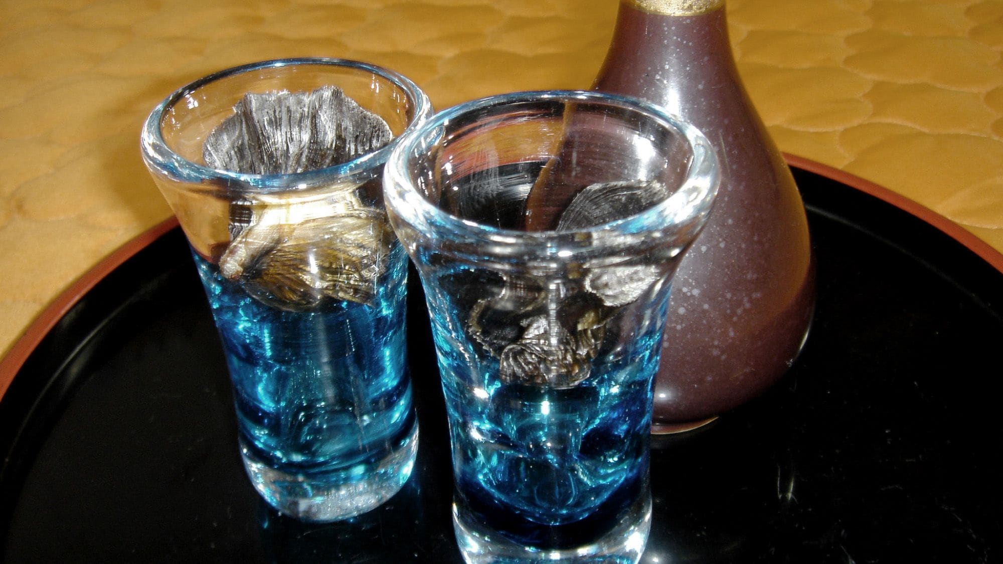 Two glasses of blue liquid on a black tray with a brown bottle.