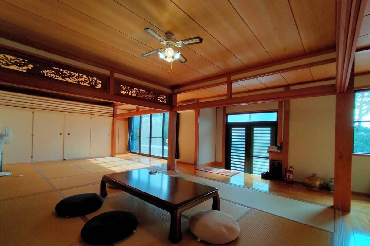 Una habitación japonesa tradicional con suelo de tatami, una mesita baja, cojines, puertas correderas y un ventilador de techo.
