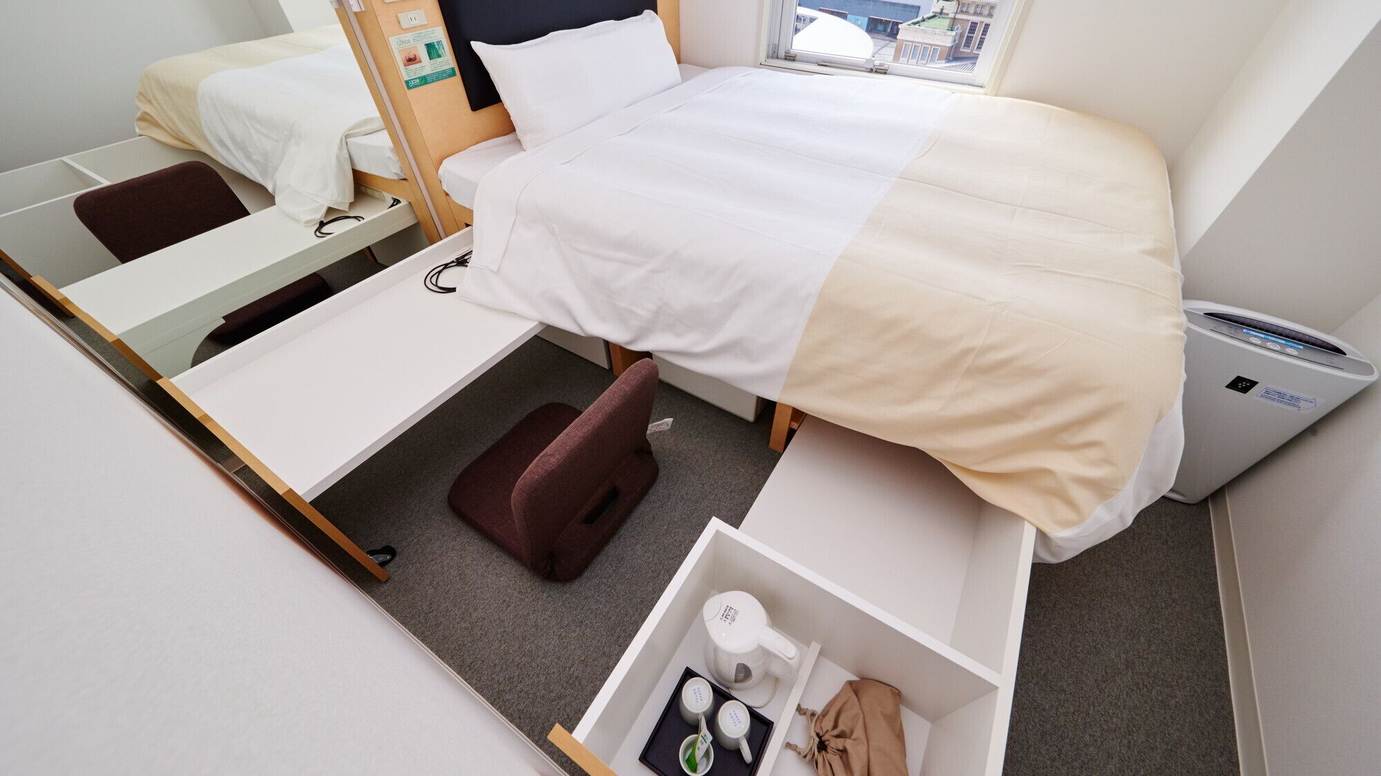 A compact hotel room with a bed, a desk, a chair, a small storage unit, and a window with a view of buildings.
