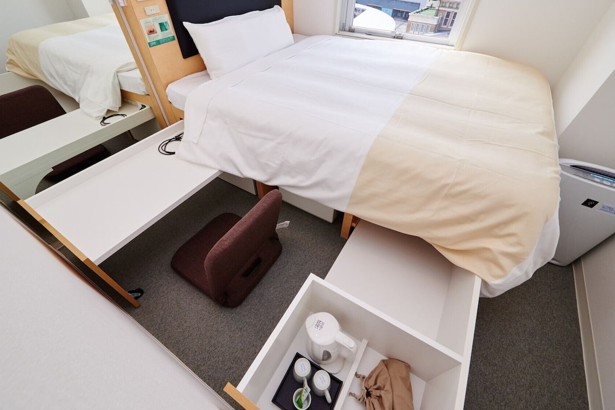 Ein kompaktes Hotelzimmer mit Bett, Schreibtisch, Stuhl, kleiner Ablage und einem Fenster mit Blick auf Gebäude.