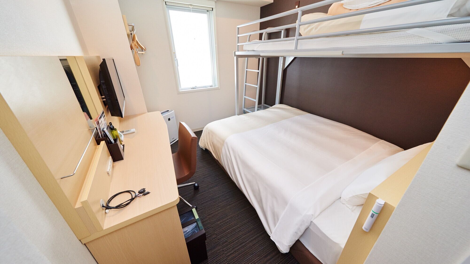 A compact hotel room with a bunk bed, a desk with a chair, a small window, and a wall-mounted TV.