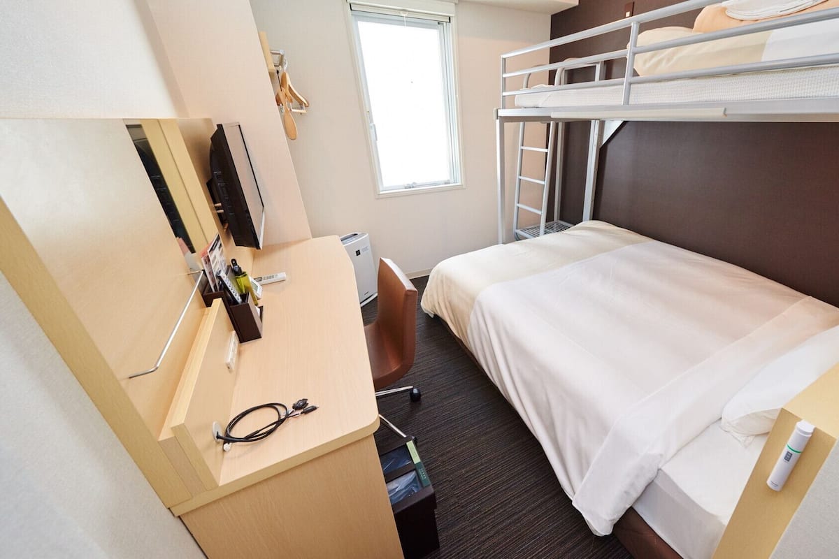 Ein kleines Hotelzimmer mit Etagenbett, Schreibtisch mit Stuhl, kleinem Fenster und Wand-Fernseher.