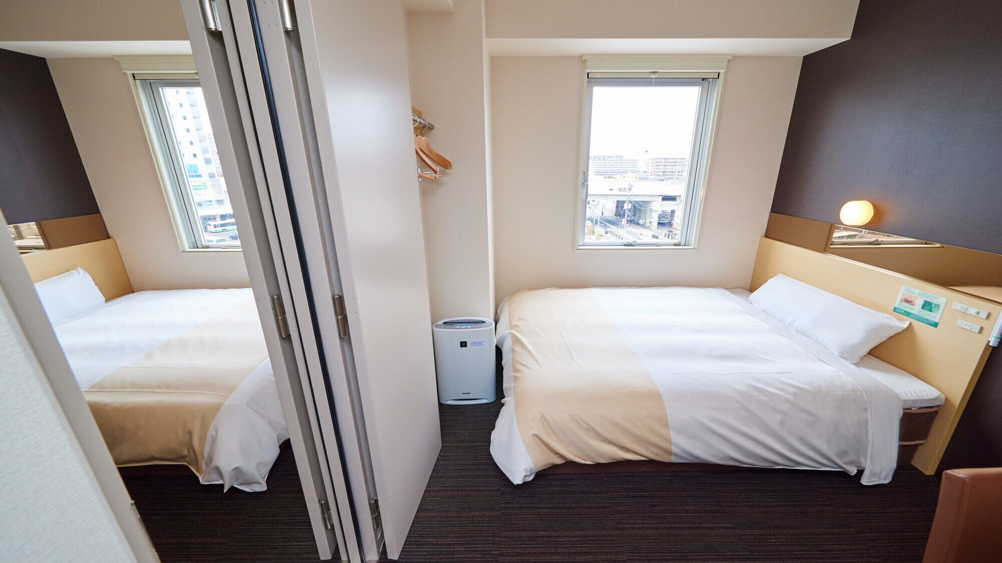 A hotel room with two beds, a window with a city view, a small trash bin, and a bedside table with a lamp.