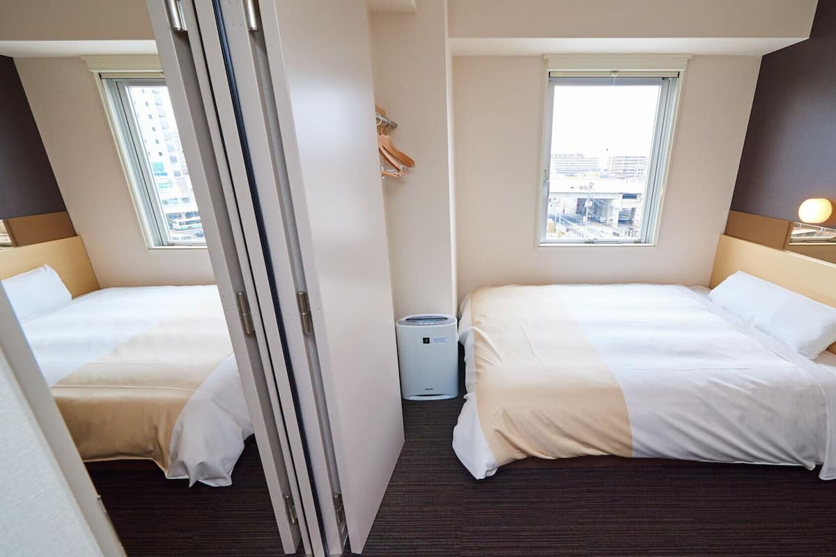 Ein Hotelzimmer mit zwei Betten, einem Fenster mit Blick auf die Stadt, einer kleinen Mülltonne und einem Nachttisch mit Lampe.
